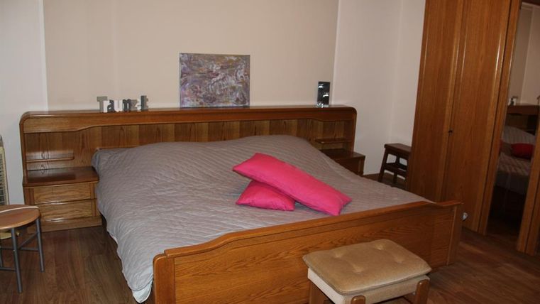 A cozy bedroom with a large wooden bed and purple pillows. The walls are in neutral colors, and there are some wooden furniture pieces.