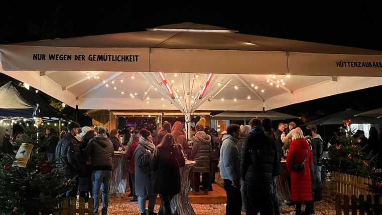 Menschen stehen an Stehtischen zusammen, im Hintergrund steht eine Hütte mit weihnachtlicher beleuchtung