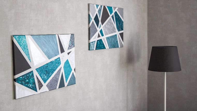 Two modern wall pictures with geometric patterns in shades of blue and gray. An elegant floor lamp stands next to it and complements the decor.