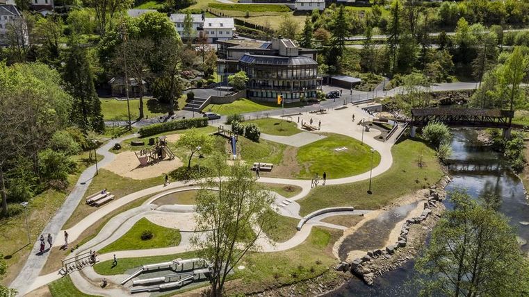 Luftaufnahme eines Kurparks mit Spielplatz, geschwungenen Wegen und einem Fluss. Im Hintergrund sind Gebäude und Bäume zu sehen.