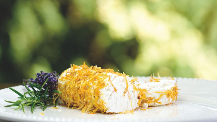 Ein leckeres Gericht mit frischem Käse, umhüllt von knusprigen Fäden, serviert auf einem Teller. Daneben liegen Kräuter und Blumen zur Dekoration.