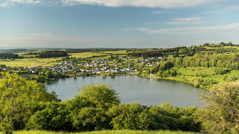 Ein malerischer Blick auf einen ruhigen See umgeben von üppigem Grün. Im Hintergrund ist ein kleines Dorf mit weißen Häusern sichtbar.