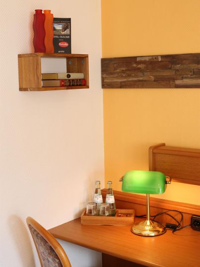 Ein gemütlicher Schreibtisch in einem Raum mit gelben Wänden. Auf dem Tisch steht eine grüne Lampe, Wasserflaschen und dekorative Gegenstände.
