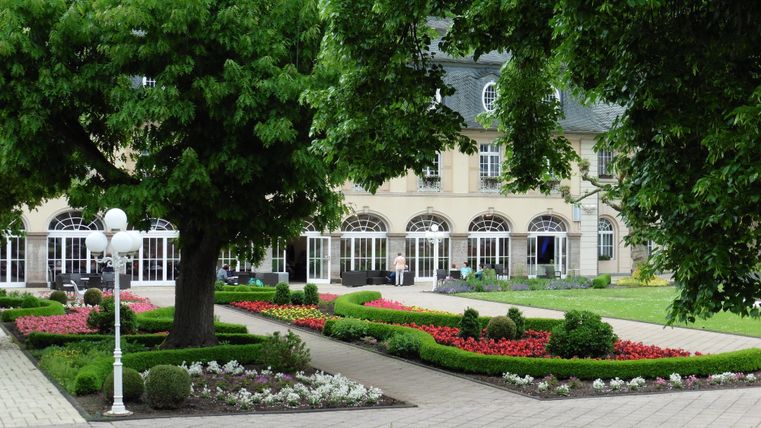 Ein gepflegter Garten mit bunten Blumen und großen Bäumen. Im Hintergrund ist ein elegantes Gebäude mit vielen Fenstern zu sehen.