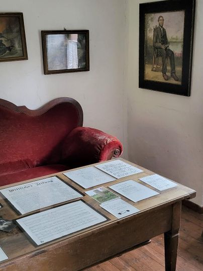 A cozy room with a red sofa and a wooden table. Documents are lying on the table, and a portrait is hanging on the wall.