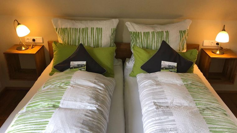 Ein gemütliches Schlafzimmer mit zwei Betten und hellen Kissen in Grün und Weiß. Neben den Betten stehen Nachtlichter und kleine Holzregale.