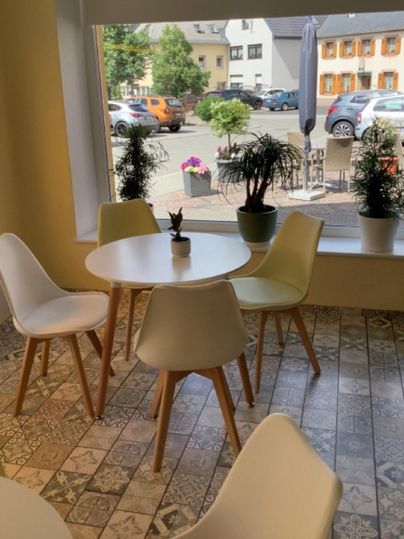 A bright café with a round table and modern chairs. Plants and parking spaces are visible through the window.