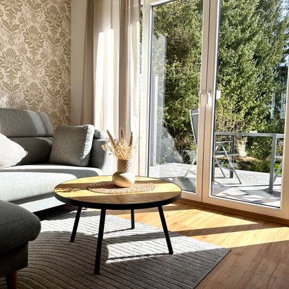 Ein gemütliches Wohnzimmer mit einem grauen Sofa und einem runden Tisch. Durch die großen Fenster sieht man einen schönen Außenbereich mit Gartenmöbeln.