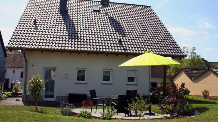 A modern house with a gabled roof and a sunny terrace. A yellow parasol provides shade on the lawn.