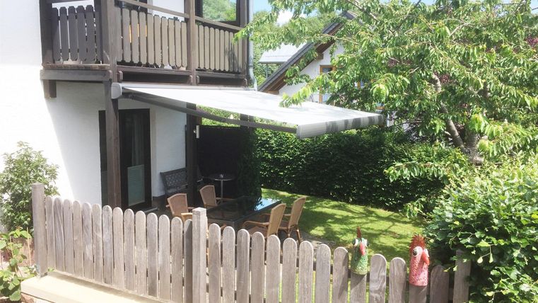 Eine gemütliche Terrasse mit einem Markisenüberdachung und Gartenmöbeln. Umgeben von grünen Pflanzen und einem Holzzaun.