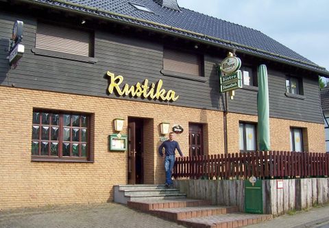 A rustic restaurant with the sign "Rustika" on the facade. In front of the entrance area, a person is standing on the steps.