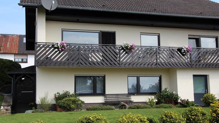 A cozy house with a balcony decorated with flowers. The garden is well-kept and lush with shrubs and plants.