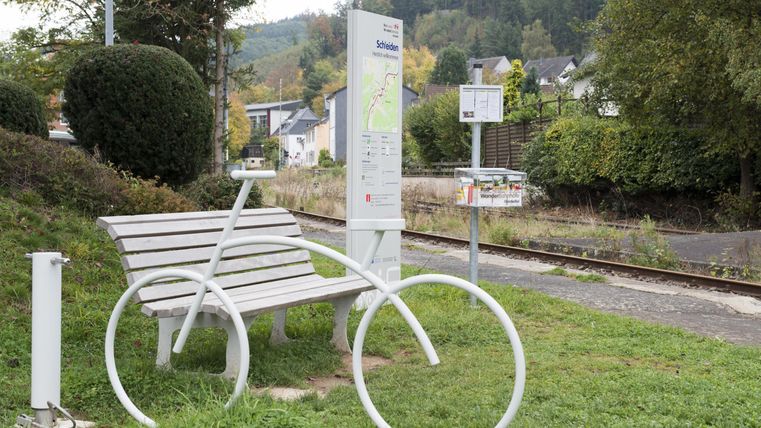 A creative bench in the shape of a bicycle stands at a bus stop. In the background, gentle hills and trees can be seen.