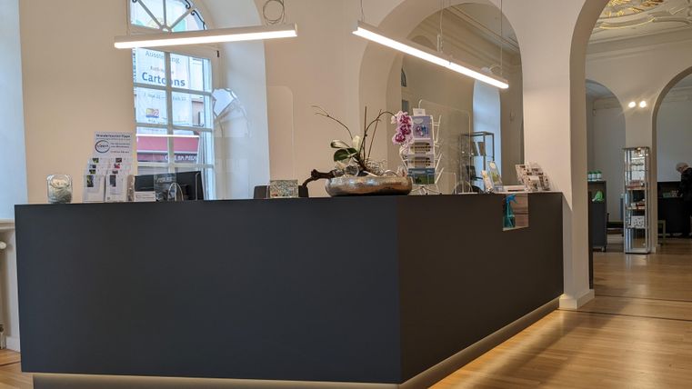 A modern reception area with a dark reception desk and warm lighting. There are some plants and information materials visible.