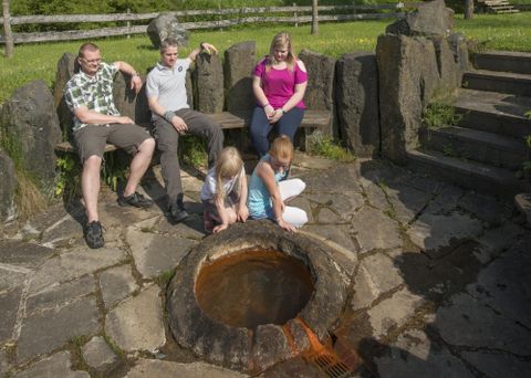 Eine Familie sitzt an einem offenen Feuerplatz aus Steinen. Kinder spielen in der Nähe des Feuers, während Erwachsene entspannt zusehen.