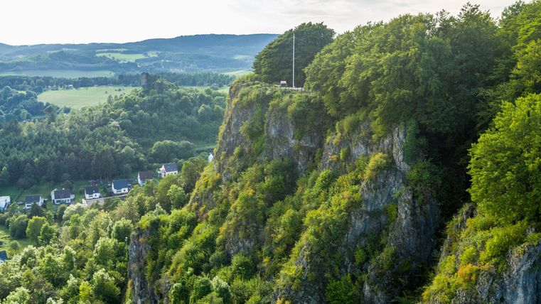 Luftaufnahme von bewaldeten Felsen mit einem kleinen Dorf im Tal. Im Hintergrund erstrecken sich grüne Hügel unter einem bewölkten Himmel.