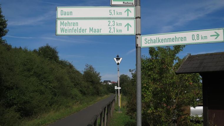Een wegwijzer toont de richtingen naar verschillende locaties: Daun, Mehrenh, Weinfelder Maar en Schalkenmehrn. De omgeving is groen en vredig, ideaal voor fietsers.