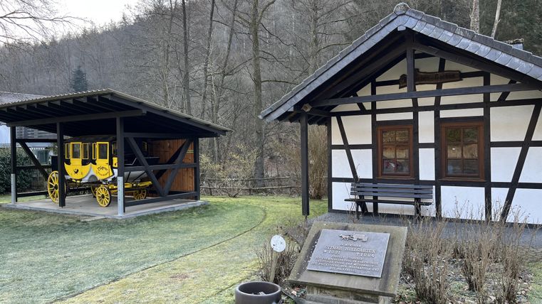 A historic building with a wooden frame and large windows stands next to a covered area where a yellow carriage is displayed. The surroundings are surrounded by trees, and a green meadow is visible.