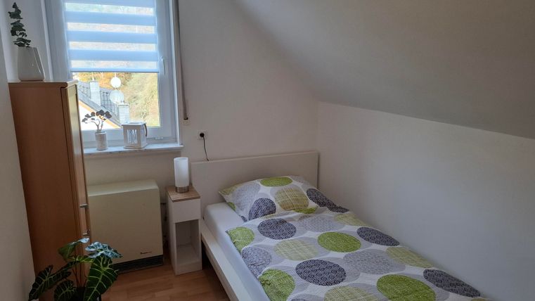 A small, bright bedroom with a single bed and colorful cushions. Next to the bed is a bedside table and a shelf, and a window allows for daylight.
