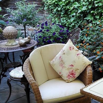 Ein gemütlicher Garten mit einem geflochtenen Sessel und einem dekorativen Kissen. Umgeben von bunten Blumen und Pflanzen.