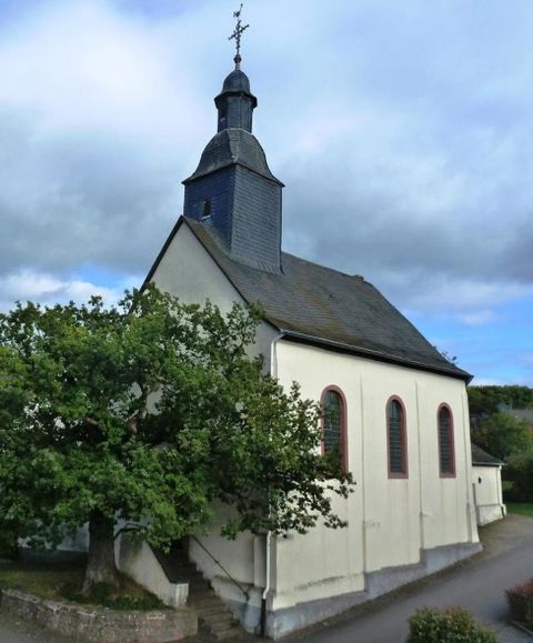 Eine kleine Kirche mit einem grauen Turm und großen Fenstern. Im Vordergrund steht ein grüner Baum.