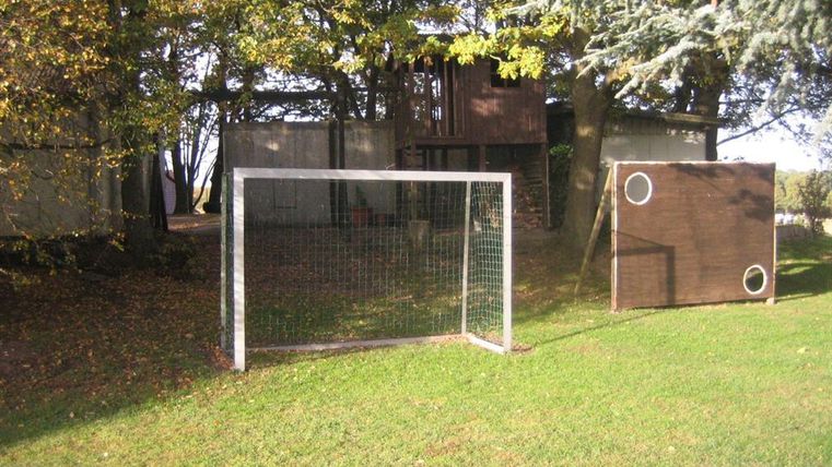 Een voetbalgoal op een groene weide, omringd door bomen. Op de achtergrond is een speelhuis zichtbaar.