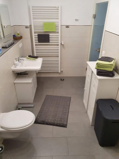 Ein modernes Badezimmer mit einem Waschbecken, WC und einem Handtuchhalter. Die Wände sind hell gefliest, und es gibt frische grüne Handtücher auf den Ablagen.