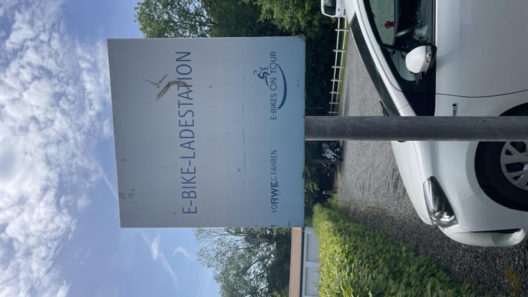 Ein Schild für eine E-Bike-Ladestation steht an einem ruhigen Ort. Im Hintergrund sind Bäume und ein Parkplatz sichtbar.