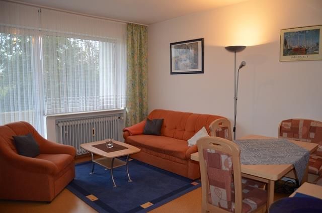 A cozy living room with two orange sofas and a table. The windows are covered with curtains and the walls are decorated with pictures.