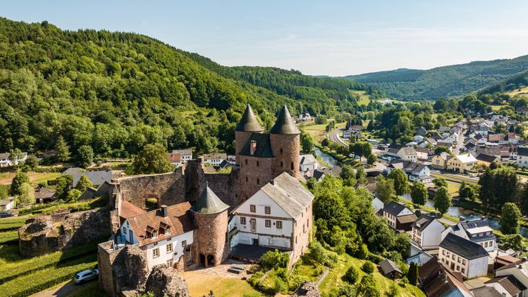 Luchtfoto van de Bertradaburg in Mürlenbach, omgeven door groene heuvels en het dorp. De burcht heeft kenmerkende torens en ligt in een idyllisch landschap.