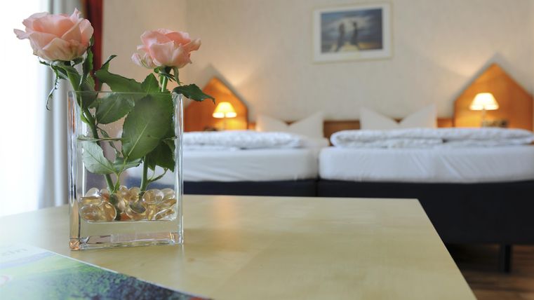 Ein gemütliches Zimmer mit zwei Betten und sanften Beleuchtungen. Auf dem Tisch steht eine Vase mit rosa Rosen.