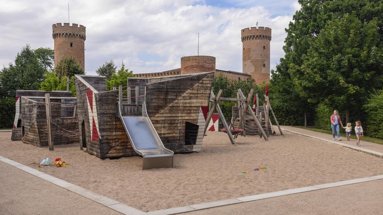 Ein Spielplatz mit einer Holzkonstruktion, Rutschen und Schaukeln. Im Hintergrund sieht man zwei Türme und viel grünes Laub.