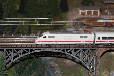 Ein Modellzug der ICE fährt über eine Brücke. Im Hintergrund sind Bäume und eine kleine Eisenbahnanlage zu sehen.