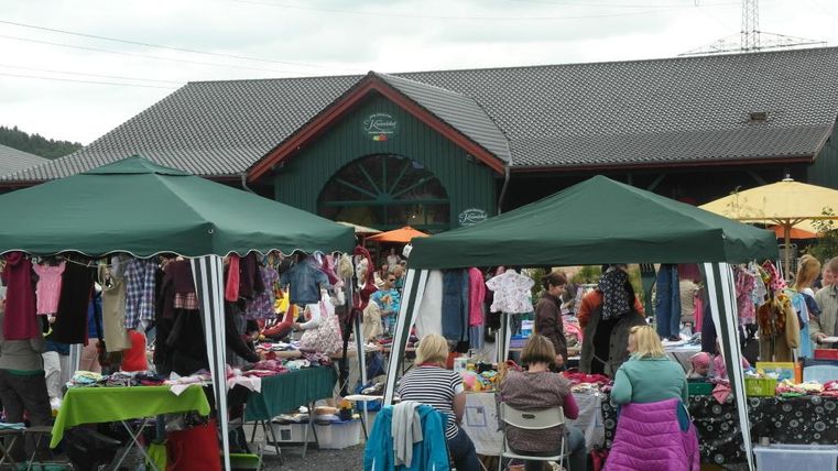 Een drukke markt met meerdere kramen en tenten. Mensen verkopen en kopen kleding en andere goederen.