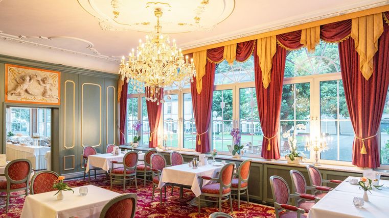 Een elegant restaurant met decoratieve rode gordijnen en een grote kroonluchter. De tafels zijn smaakvol gedekt en bieden uitzicht op het terras.