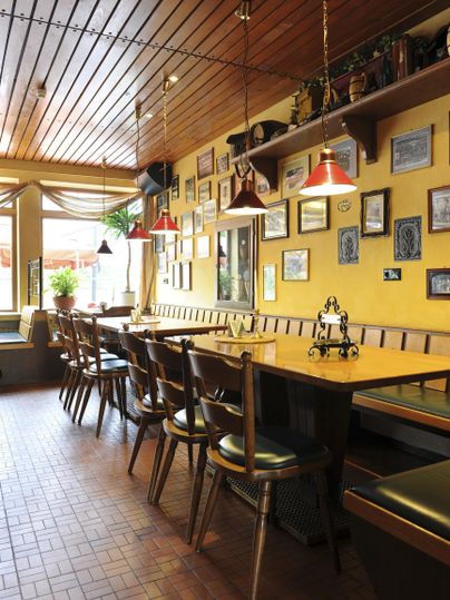 Ein gemütliches Restaurant mit Holzwänden und einer langen Tischreihe. An den Wänden hängen verschiedene Bilder und die Beleuchtung ist warm und einladend.