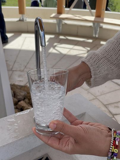 Een persoon vult een glas met water uit een kraan in een buitenruimte. Op de achtergrond zijn mensen te zien.