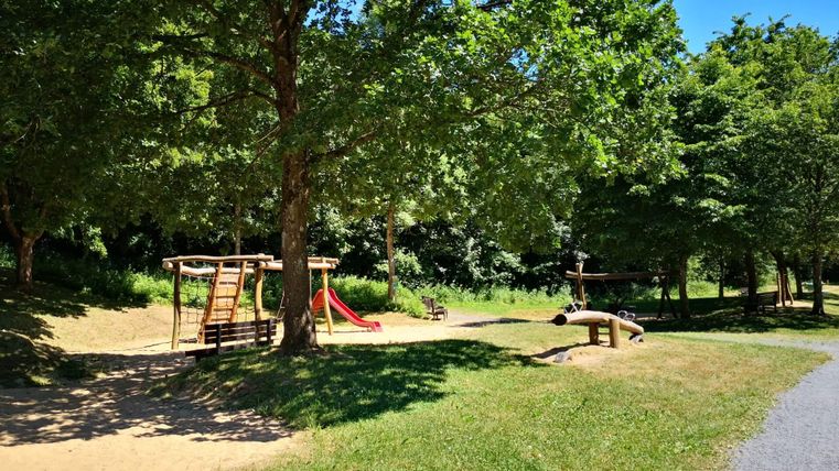 Een speelplaats met een glijbaan en een klimrek onder grote bomen. De omgeving is groen en uitnodigend.