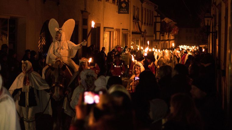 A crowd is celebrating on a festive night with torches. In the foreground, there is a group of people in costumes.
