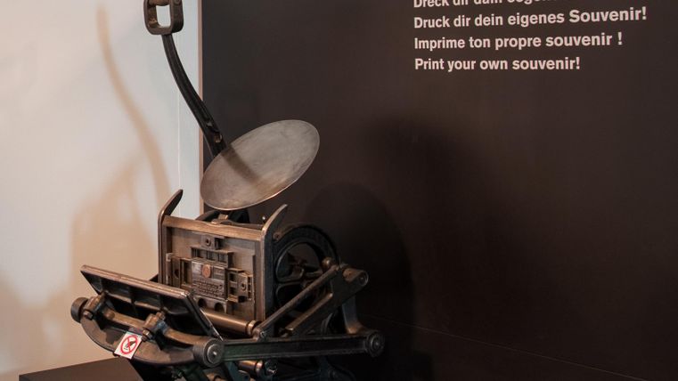 An old printing press is standing on a table. Next to it is a wall with a note stating that you can print your own souvenir.
