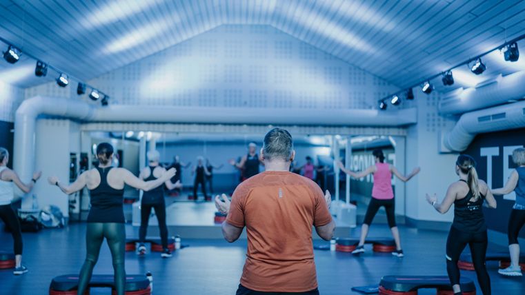 Een groep mensen traint in een moderne sportschool. Ze doen een fitnessoefening met stepborden en worden geleid door een trainer.