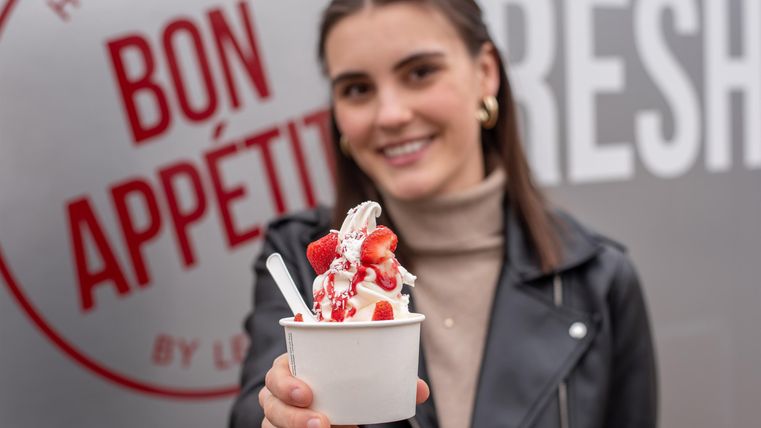 Een jonge vrouw houdt een ijscoupe met aardbeien en slagroom vast. Op de achtergrond is een herkomstlabel met de tekst "Bon Appétit" te zien.