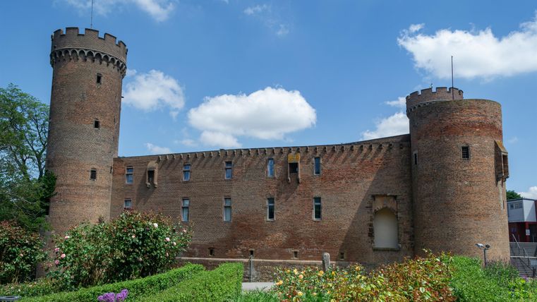 A historical castle with two towers and red brick walls. In the foreground, colorful flowers are blooming and the sky is blue with some clouds.