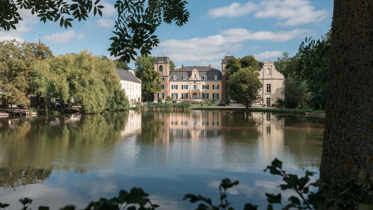 Een schilderachtig landschap met een rustige vijver en historische gebouwen op de achtergrond. De afbeelding toont een vredige sfeer met mooi weer.