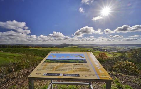 Een uitzichtpaneel op een heuvel met groene velden en een heldere lucht. De zon schijnt fel en er zijn enkele wolken zichtbaar.