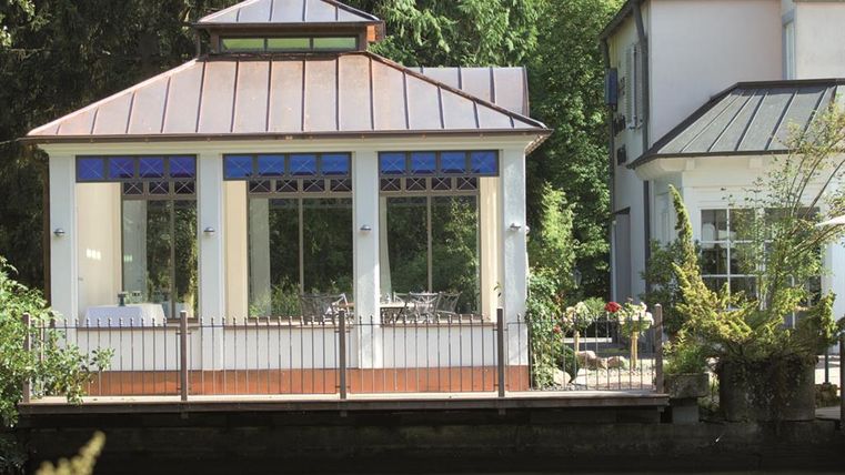 A well-maintained garden house with large windows and a covered area. It is located by a quiet body of water, surrounded by plants and trees.