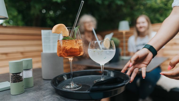 Ein Tablett mit zwei Getränken steht auf einem Tisch. Im Hintergrund sitzen lächelnde Gäste in einer gemütlichen Umgebung.