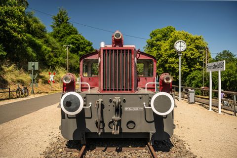 An old locomotive at the train station. In the background, trees and a clock are visible.