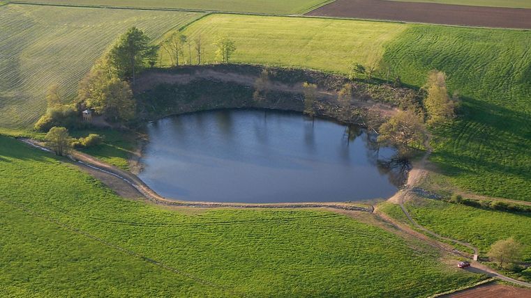 Luftaufnahme des Eichholzmaars, ein kreisförmiger See umgeben von grünen Feldern und Bäumen, mit einem kleinen Weg und einem Fahrzeug am Ufer.