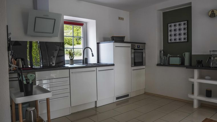 Eine moderne Küche mit weißen Möbeln und schwarzer Arbeitsplatte. Sie hat große Fenster und ist gut beleuchtet.